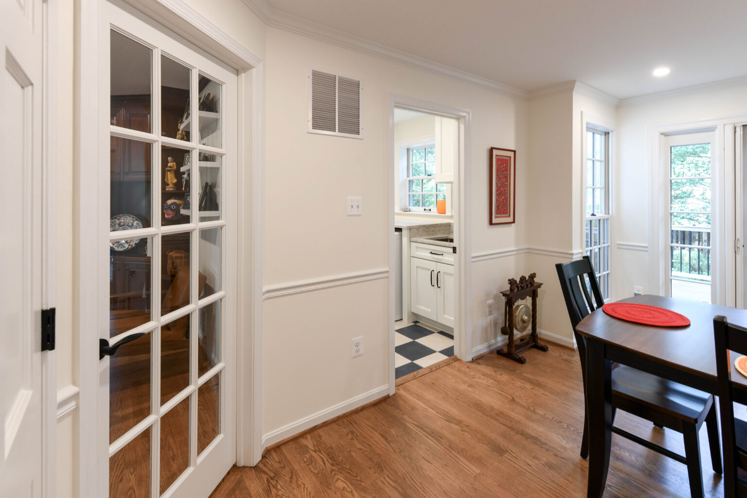 A dining area with a wooden table, glass door to a pantry, view of a kitchen with checkered floor, white walls, and wood floors throughout.