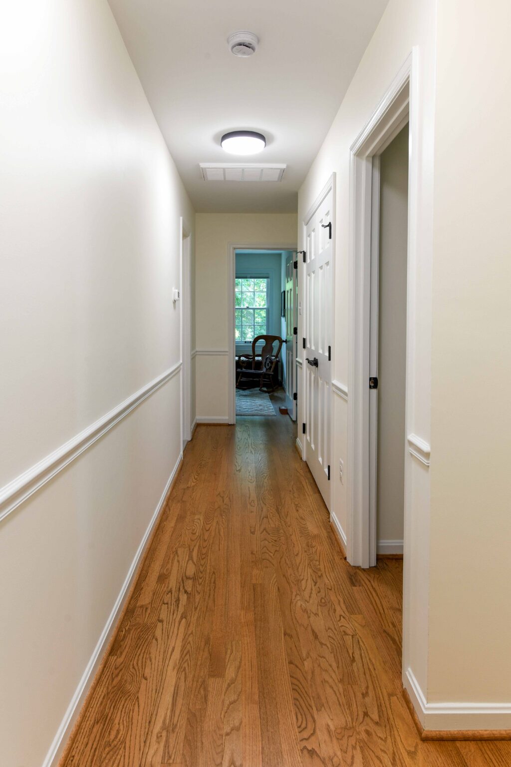 A narrow hallway with light-colored walls and wood flooring leads to an open door, revealing a room with a window and a chair.