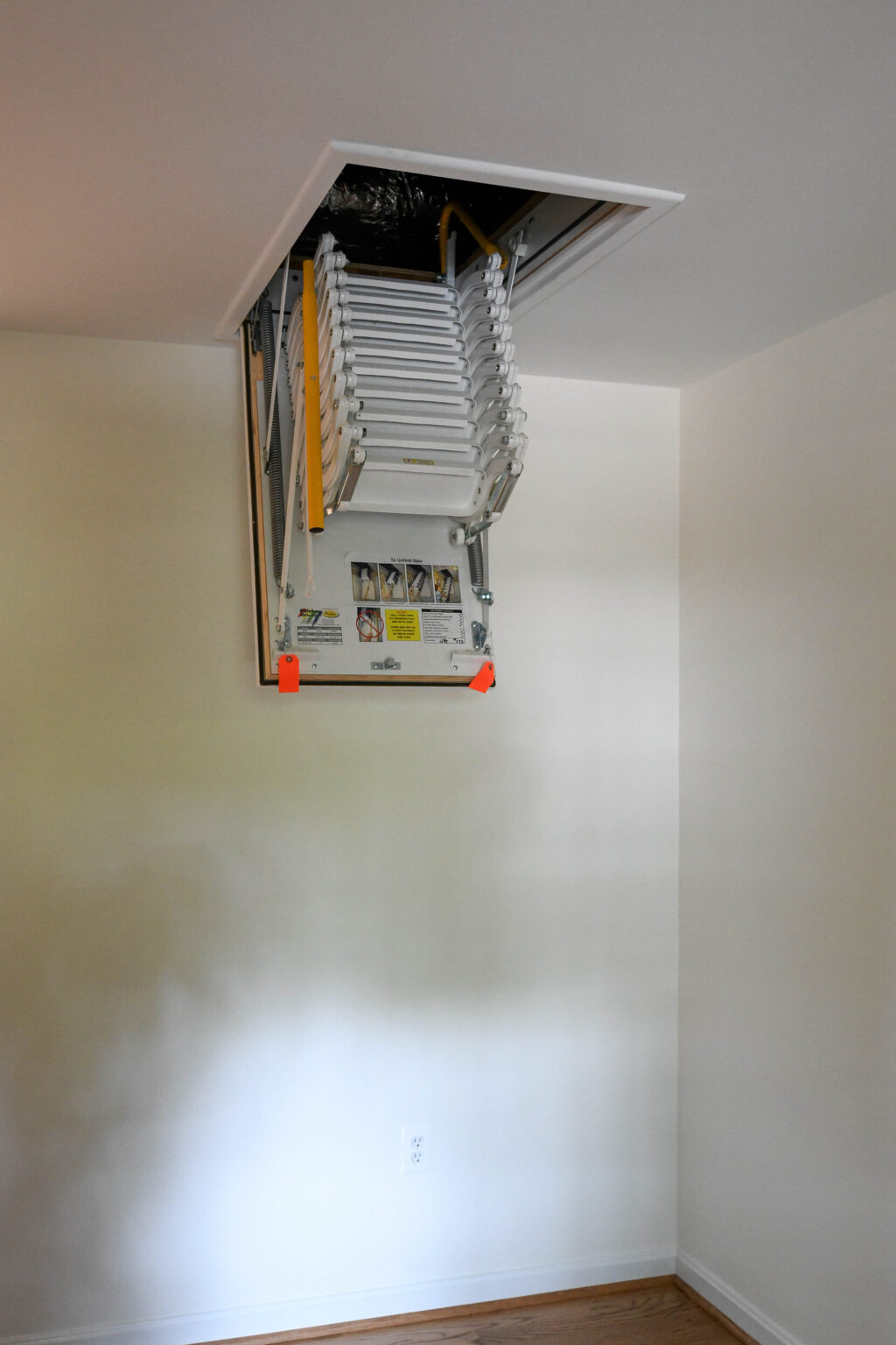 Folding attic ladder partially extended from a ceiling hatch in a white room with hardwood flooring.