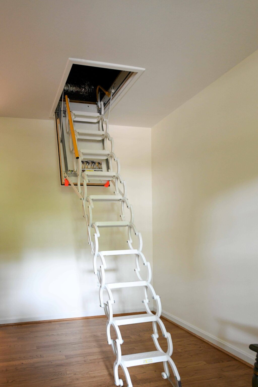 Foldable attic ladder extended from a ceiling hatch, with a yellow handrail and a wooden floor visible below.