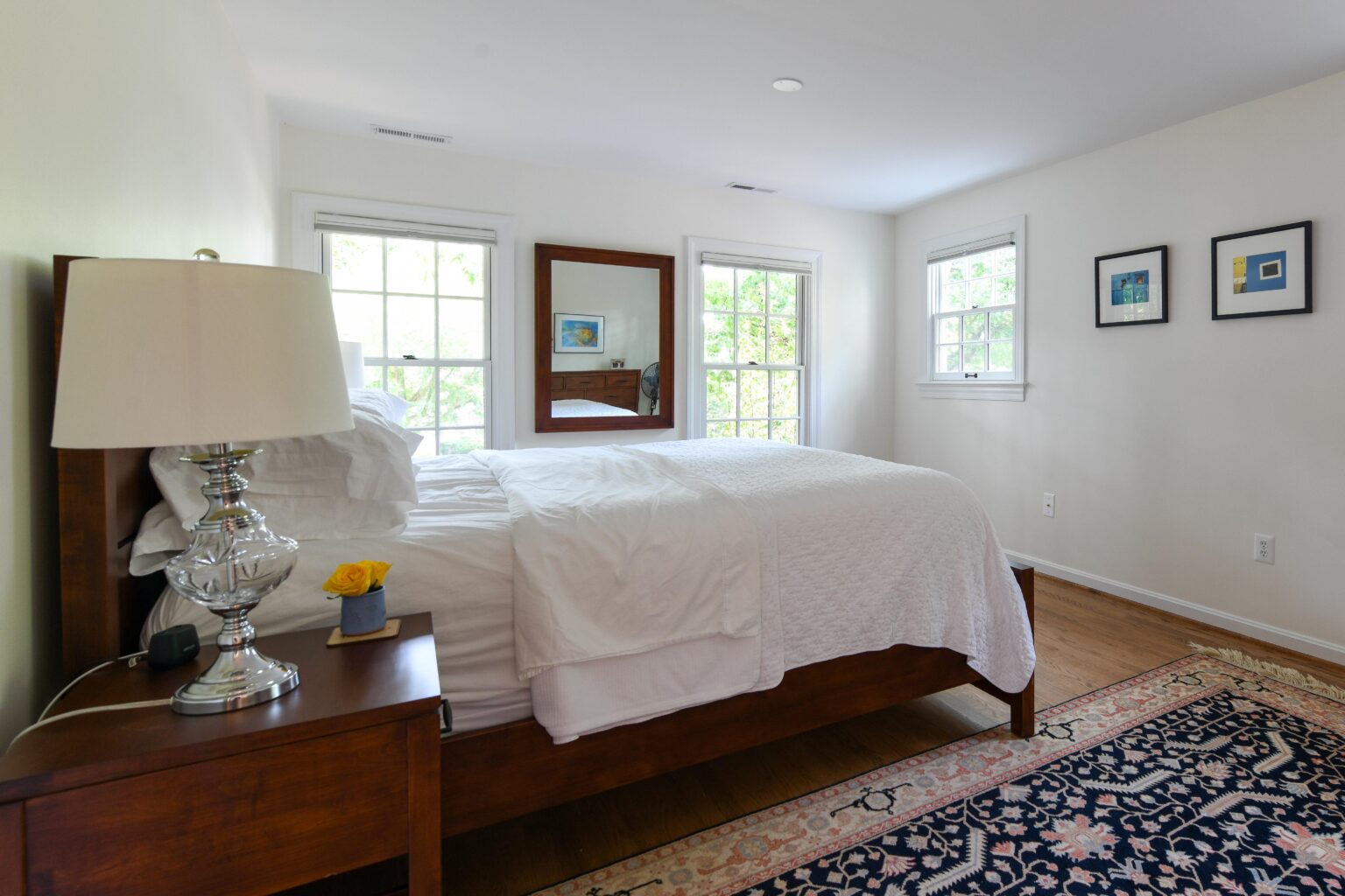 A neatly made bed with white bedding sits beside a wooden nightstand and lamp in a bright bedroom with windows, framed art, and a patterned rug.
