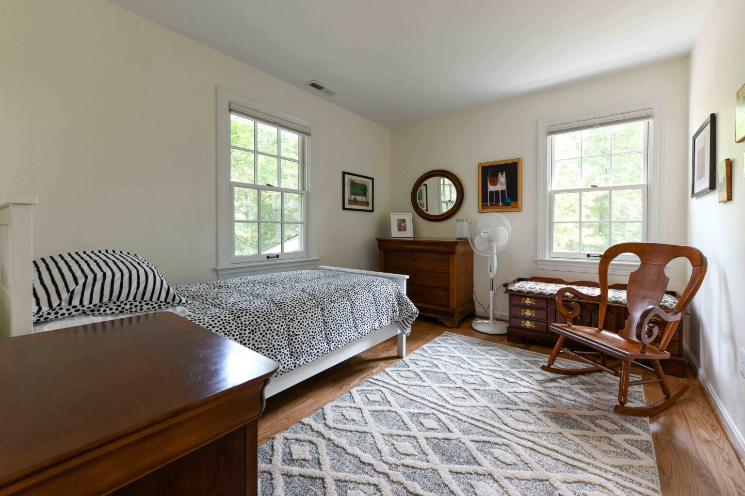 A bedroom with a single bed, wooden furniture, a patterned rug, a fan, and three windows letting in natural light.