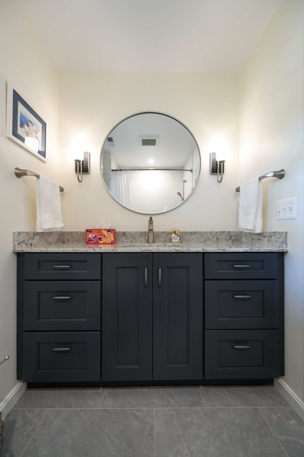 Modern bathroom vanity with dark cabinets, marble countertop, round mirror, two wall sconces, hand towels on racks, and framed art on a light-colored wall.