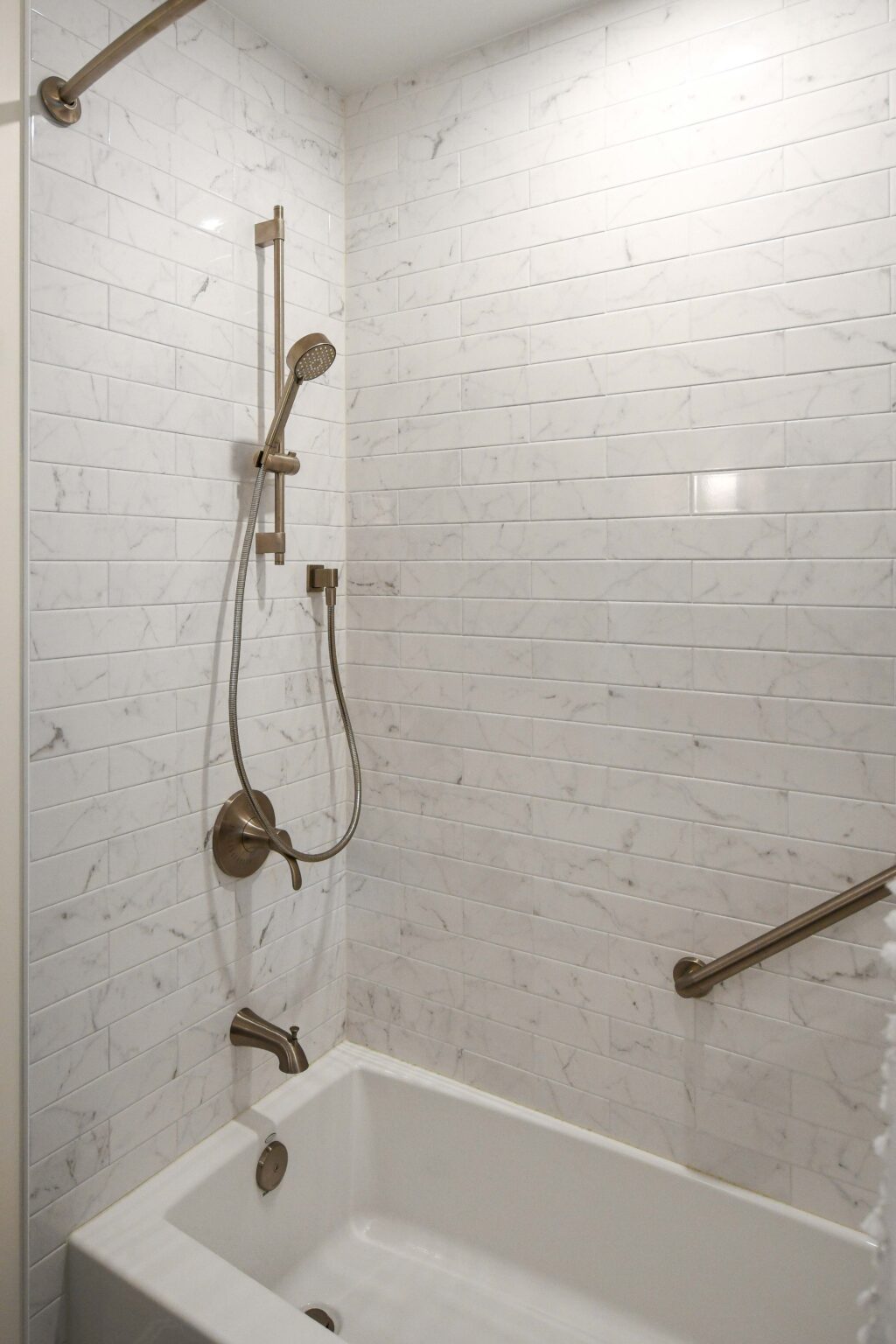 Modern bathroom with a white marble-tiled wall, a showerhead with flexible hose, a bathtub, grab bar, faucet, and control handle.