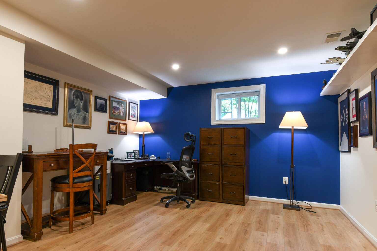 A home office with wood floors, a blue accent wall, a desk with a chair, a filing cabinet, floor lamp, and framed art on the walls. A small window lets in natural light.