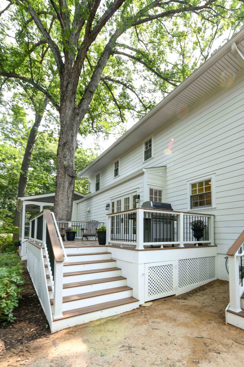 Back view of a two-story white house with a wooden deck, stairs, black railings, potted plants, and large trees providing shade.