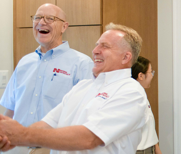 Two men in collared shirts laugh and shake hands indoors while a woman stands in the background, facing away.