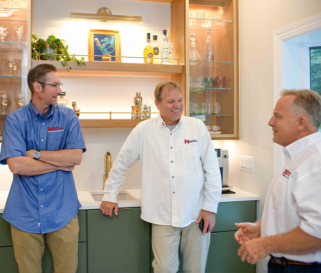 Three men in collared shirts stand and talk in a modern kitchen with glass shelves holding bottles and glassware.