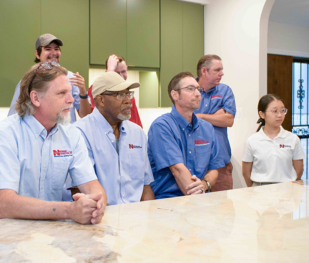 Seven people in work uniforms stand and sit around a kitchen island, looking toward something out of frame. The setting is indoors with green cabinets in the background.