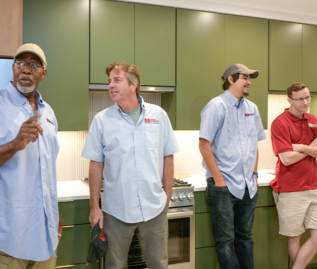 Four men stand in a modern kitchen, three wearing light blue shirts and one in a red shirt. They appear to be listening and talking.