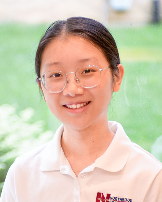 A young person with glasses and long hair tied back smiles, wearing a white polo shirt with a Northwood school logo, standing indoors with a blurred green background.