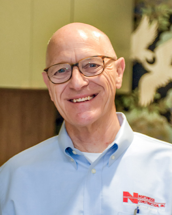 A bald man with glasses smiles, wearing a light blue button-up shirt with a red Northside Construction, Inc. logo, standing indoors.
