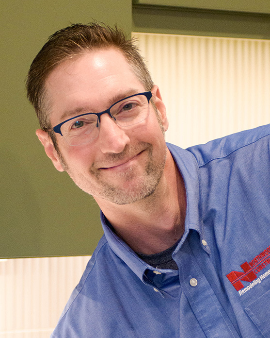 Man with short brown hair and glasses, wearing a blue collared shirt with a red embroidered logo, smiling indoors with a green cabinet in the background.
