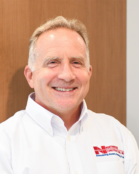 A middle-aged man with short light hair smiles while wearing a white Northwood Construction company shirt, standing in front of a wooden background.
