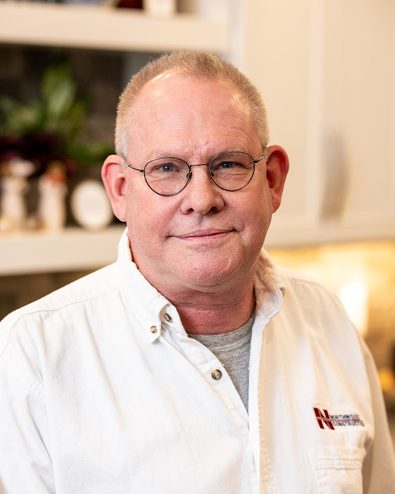 A middle-aged man with glasses and short hair, wearing a white button-up shirt and gray t-shirt, standing indoors in front of a blurred kitchen background.