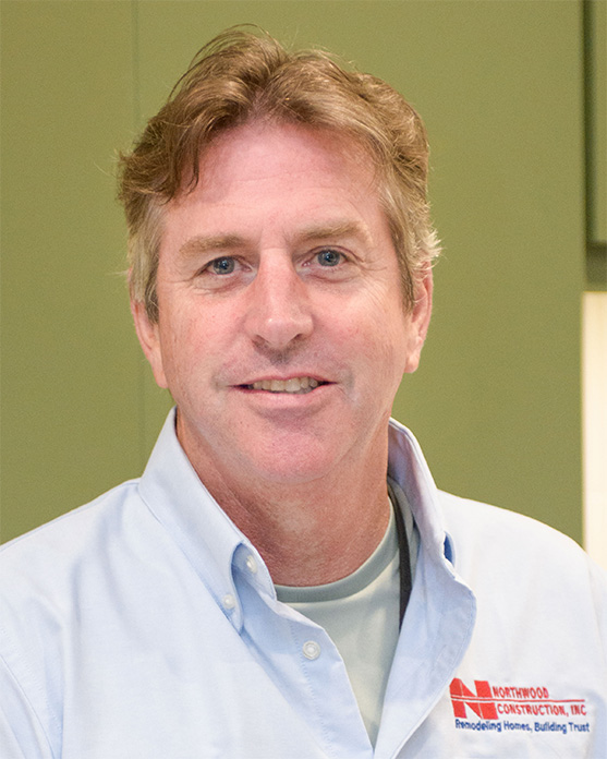 A man with light brown hair wearing a light blue collared shirt with a Northwood Construction, Inc. logo stands indoors, smiling at the camera.