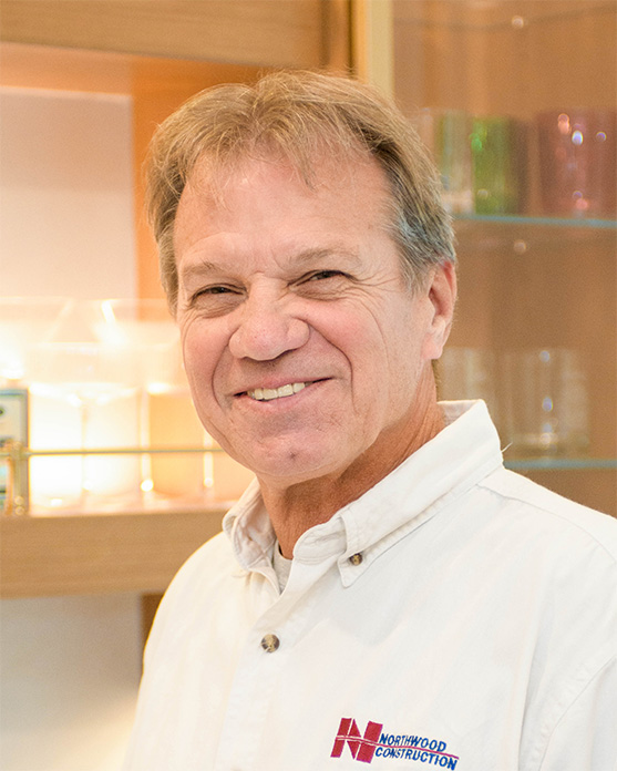 Smiling man in a white Northwood Construction shirt stands indoors in front of glass shelves with various glassware, proudly representing our team.