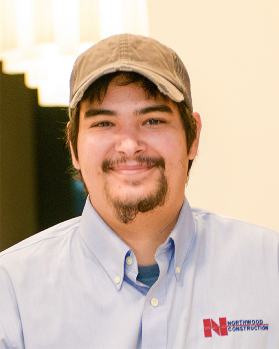 A person wearing a gray cap and light blue Northwood Construction shirt smiles at the camera indoors.