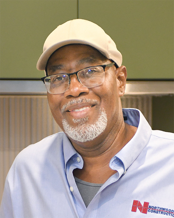 Smiling man with glasses, white beard, and beige cap, wearing a blue shirt with a Northwood Construction logo, stands indoors.