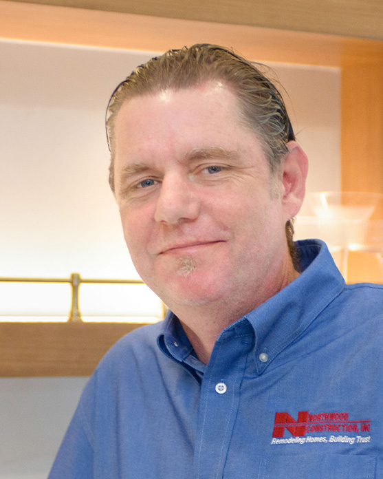 Man in a blue collared shirt with a company logo stands indoors, smiling slightly at the camera. Shelves and home decor are visible in the background.