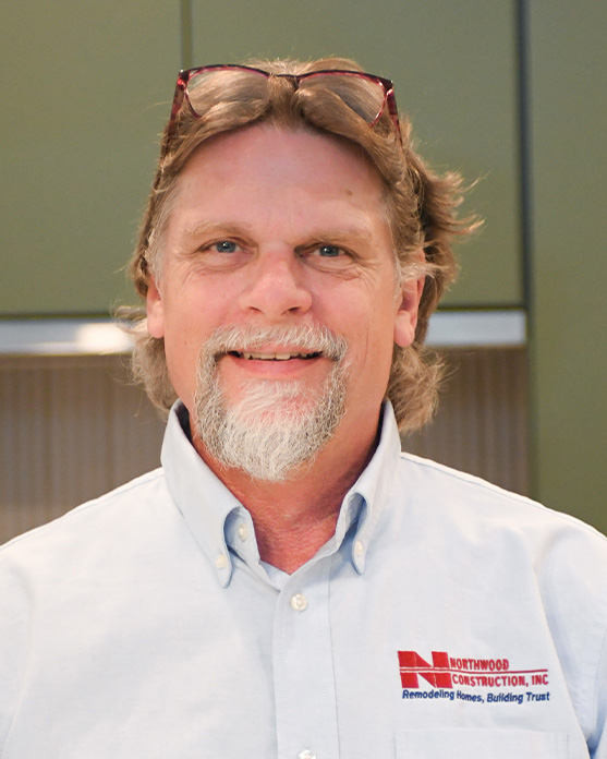 Man with light brown hair, glasses on head, and gray beard, wearing a light blue Northwood Construction, Inc. shirt, smiling indoors.