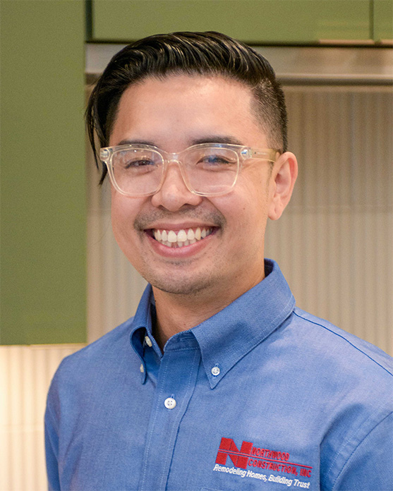 Smiling man with short dark hair, glasses, and a blue collared shirt stands indoors. The shirt has a construction company logo and slogan on it.