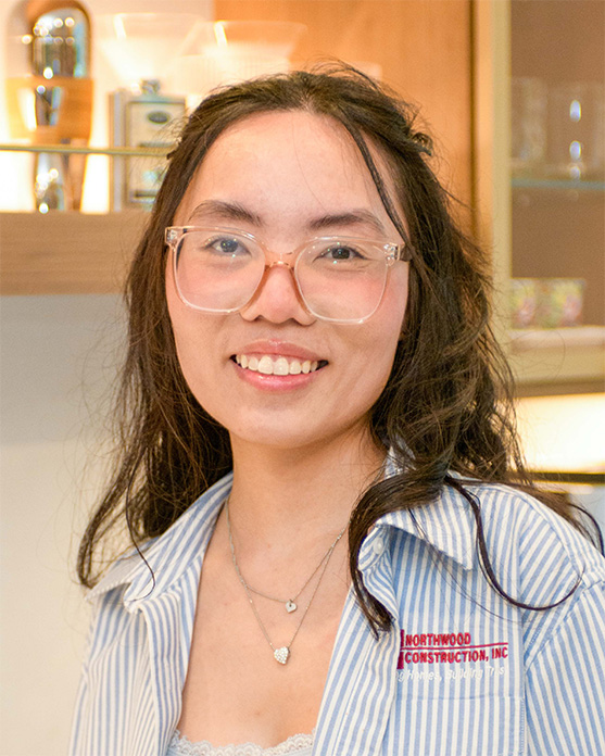 A person with long dark hair and glasses smiles at the camera, wearing a striped shirt that reads “Northwood Construction, Inc.” Kitchen items are visible in the background.