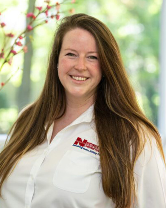 Woman with long brown hair wearing a white shirt with a red and black logo, smiling in front of a blurred, green outdoor background with pink flowers.