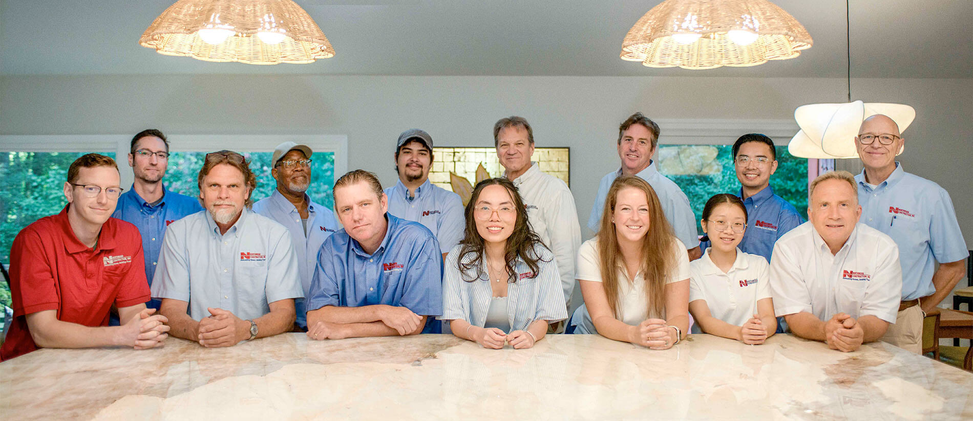 A group of fourteen people, wearing company shirts, pose together behind a marble countertop in a well-lit room with modern lighting fixtures.