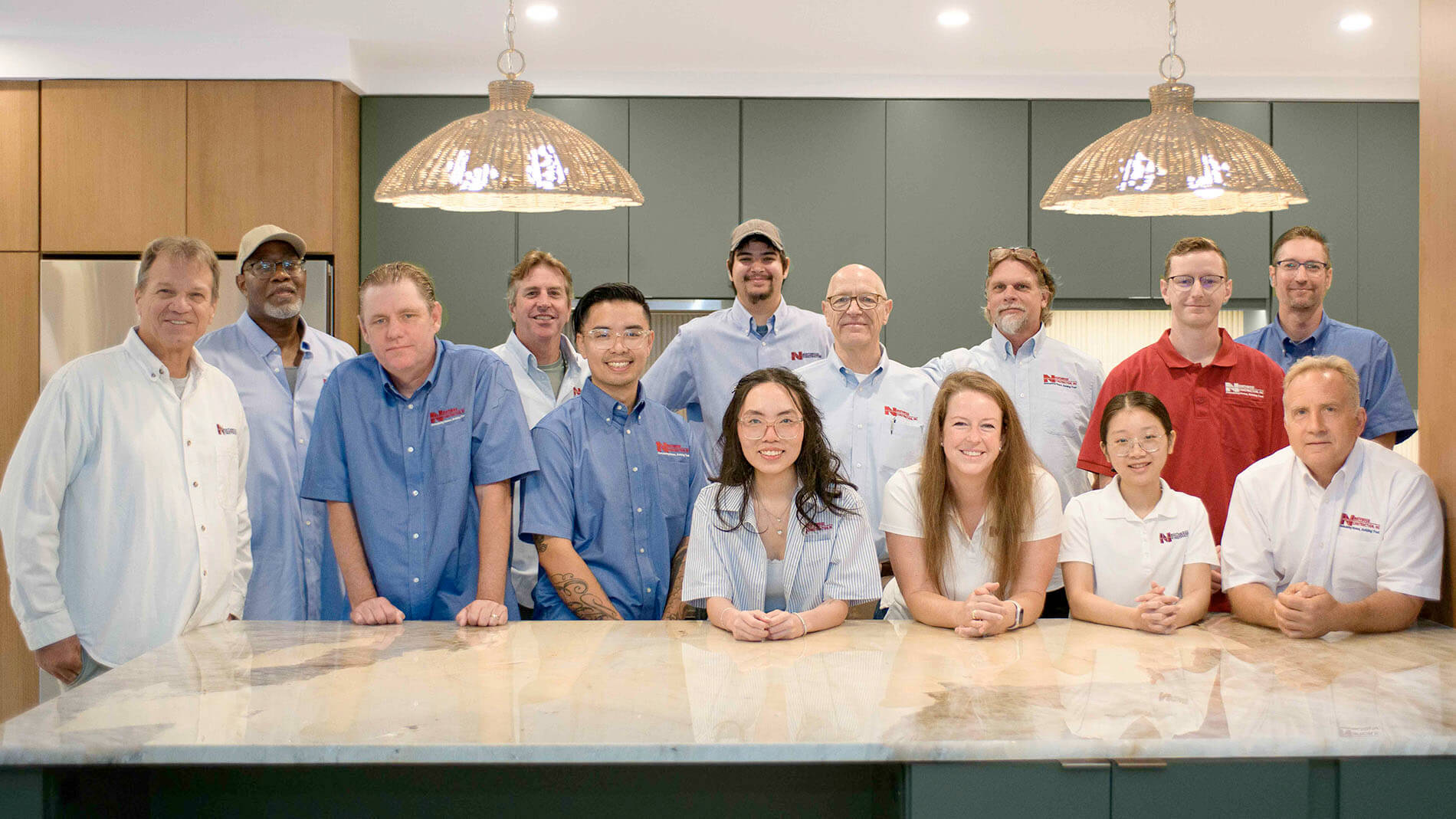 Thirteen people pose together in a modern kitchen, standing and sitting behind a marble counter under two pendant lights—a perfect snapshot of camaraderie during the home remodeling process.