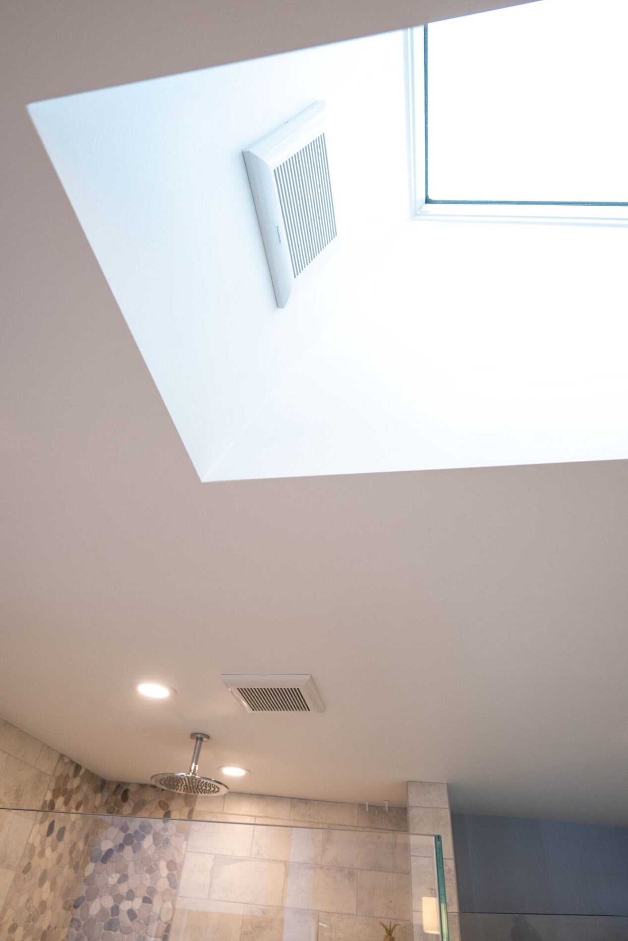 A white ceiling in a bathroom featuring a large skylight with an integrated ventilation grate, a shower head, and recessed lighting.