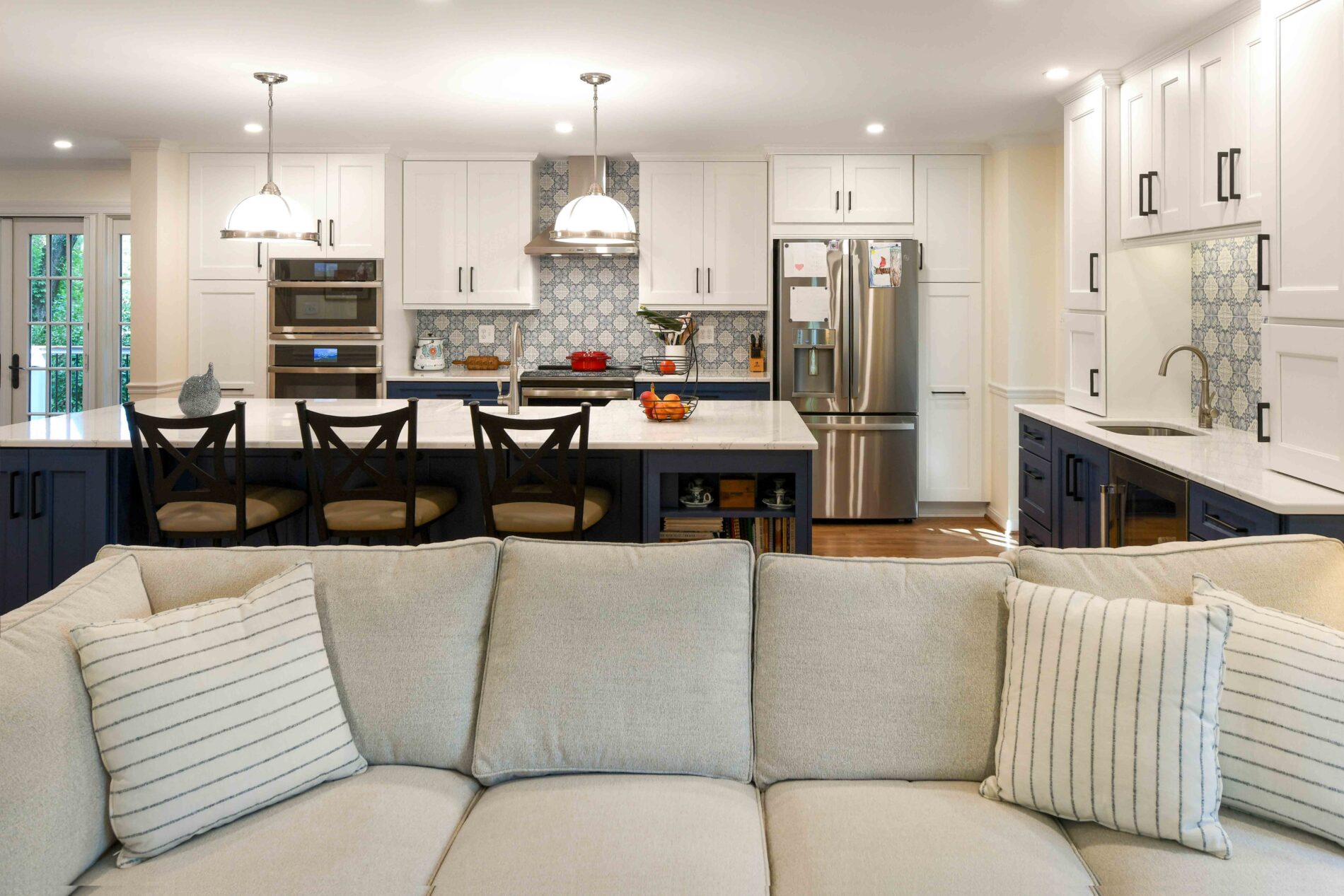 A kitchen with white cabinets, blue lower cabinets, patterned tile backsplashes, and a central island, viewed from behind a light-colored sofa.