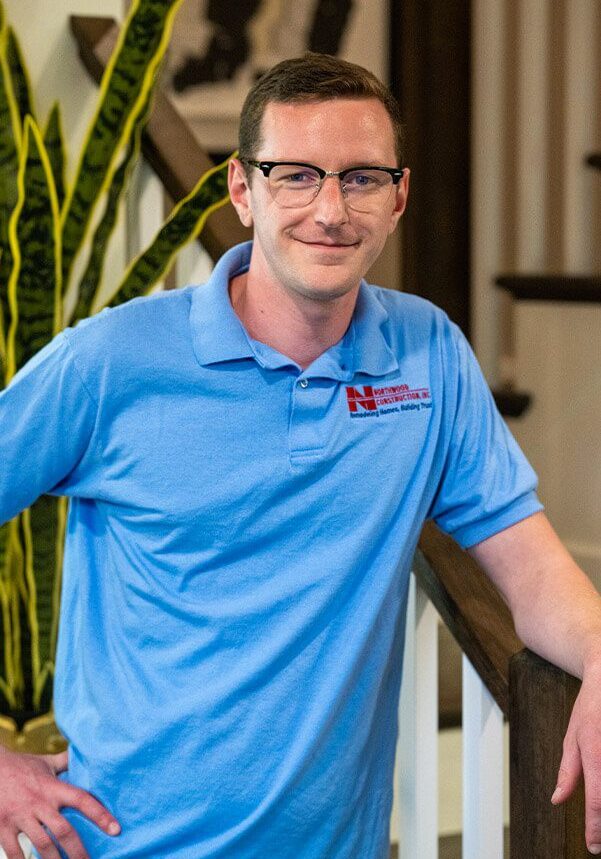 A member of our team, wearing glasses and a light blue polo shirt, stands indoors, resting his arm on a wooden railing. A tall plant is visible in the background.